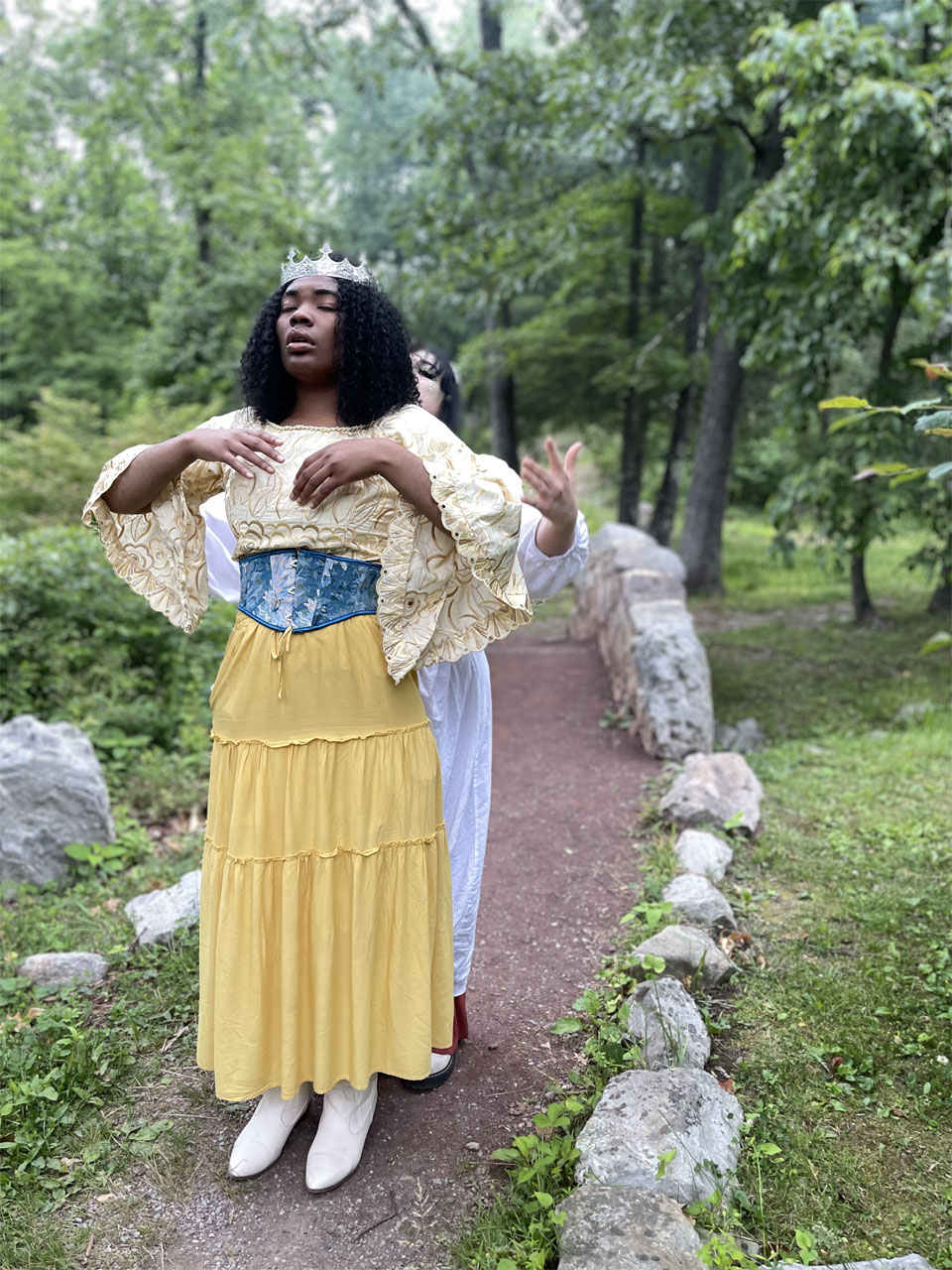 A dark-skinned young woman with curly hair, wearing a silver crown and light-colored, flowing clothes, stands on a footpath with a stone bridge and woods in the background, her eyes closed as she begins to raise her arms. Close behind her a light-skinned young woman gestures as if invoking magic to control the crowned woman.