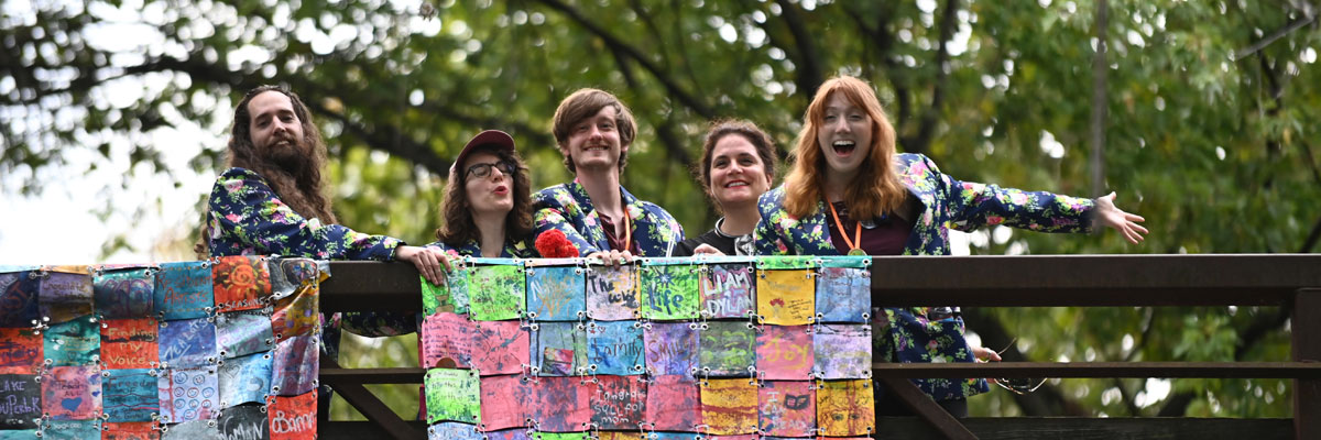 Five young adults with light skin and various gender expressions, all wearing blue jackets with a pink and green flower print, smile and wave from a bridge.