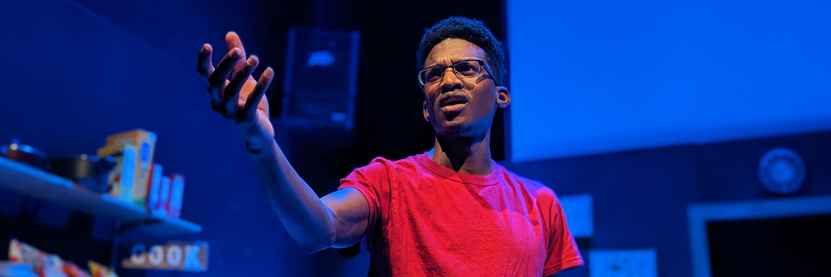 A dark-skinned young man with glasses and a red T-shirt looks up and gestures, standing in a kitchen-like area that's bathed in blue light.