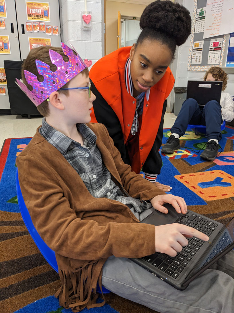 A young woman leans over an elementary-age boy wearing glasses and a pink crown as he points to show her something on his laptop screen. Behind them, another student in the classroom sits on a brightly-colored rug while working on another laptop.